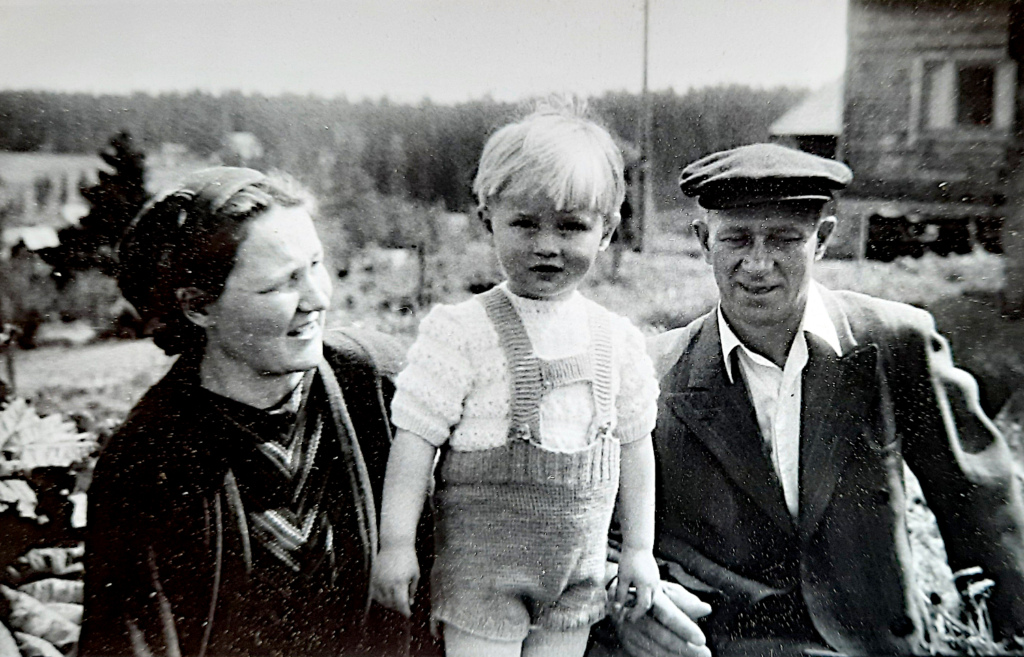 Pikkupoika vanhempineen maalaismaisemassa.