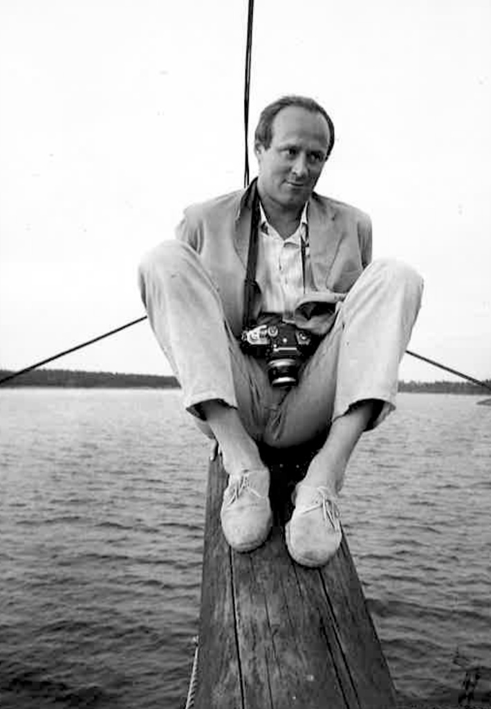 Mies veneen keulassa kamera kaulassa roikkuen.