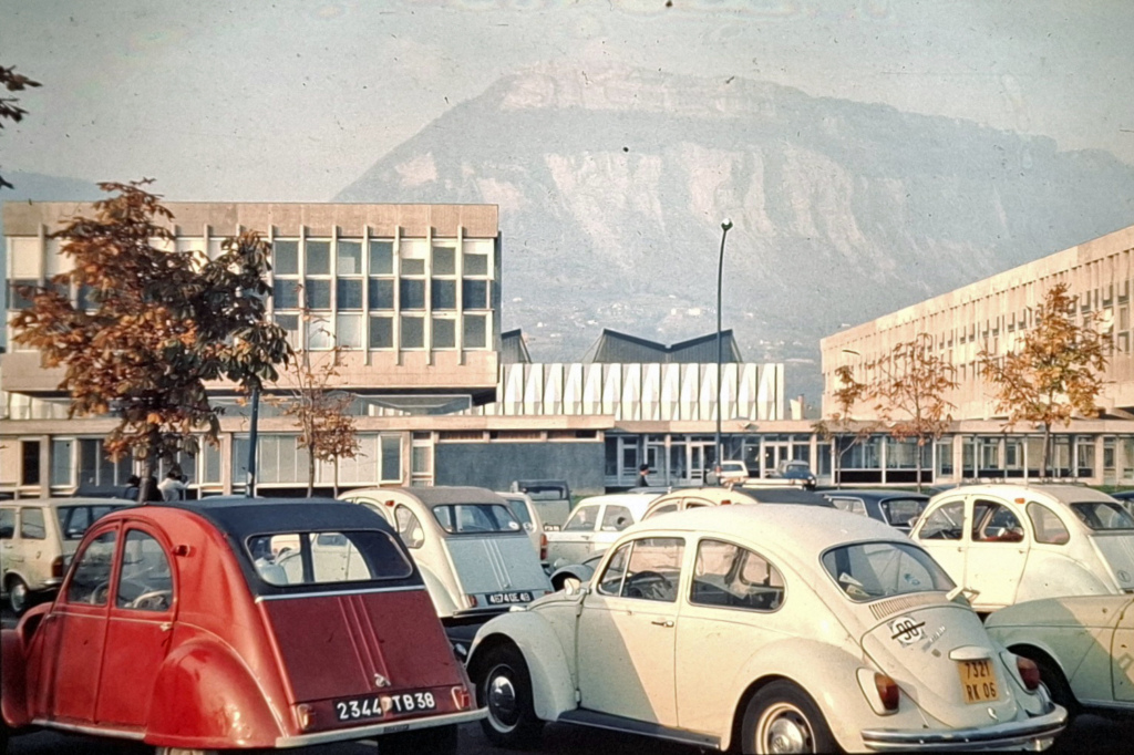 VW Kupla- ja 2CV-autoja parkissa yliopistorakennuksen edessä, taustalla vuori.