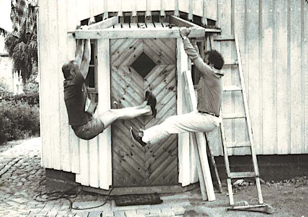 Kaksi miestä roikkuu katoksen tukilaudoissa.